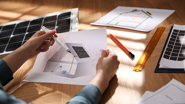 Eine Person hält einen Hausplan, auf dem am Dach eine Photovoltaikanlage und im Haus die dazugehörige Infrastruktur zu sehen ist.