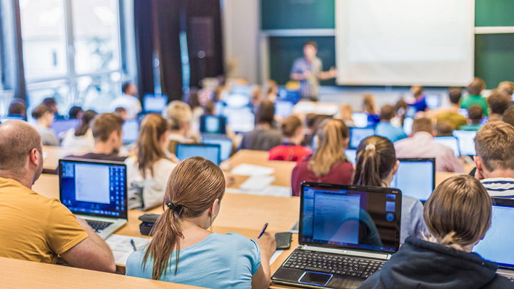 Studierende sitzen in den Reihen eines Hörsaals vor eingeschalteten Laptops und hören einem Vortragenden zu.