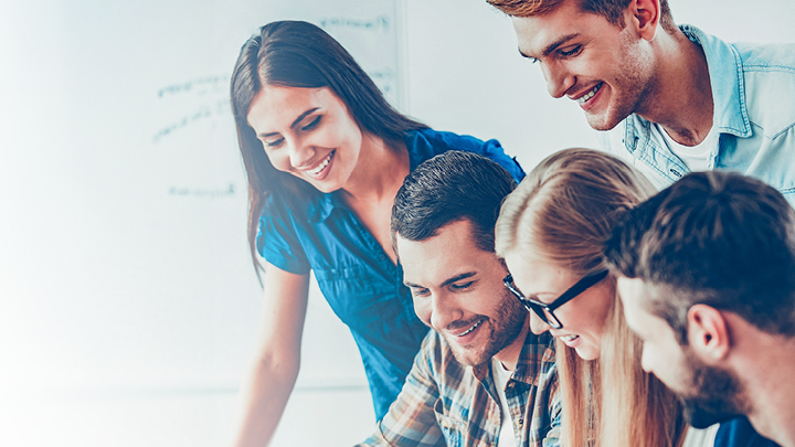 Mehrere Erwachsene blicken lächelnd auf etwas herab, wahrscheinlich versammeln sie sich um ein Lehrbuch.